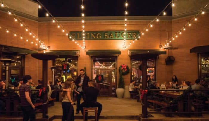 Nighttime view of a lively brick-walled restaurant patio with string lights, diners, and patio heaters.
