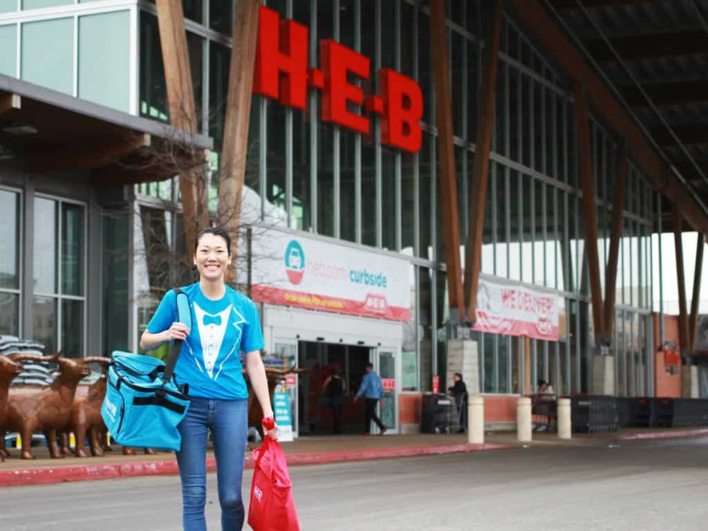 Smiling person with bags outside HEB grocery store entrance.