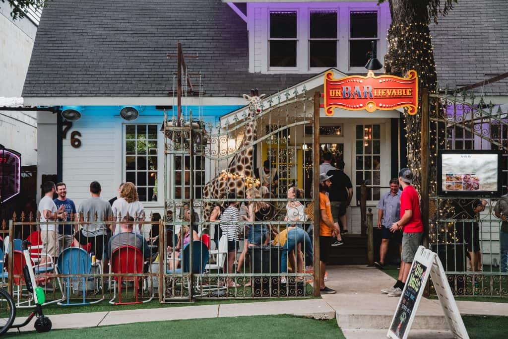 Crowd outside a lively bar with a unique giraffe decoration in front, featuring colorful seating and festive lights.