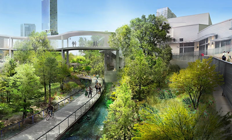 Lush urban park with trees and a walkway bridge beneath modern buildings on a sunny day.