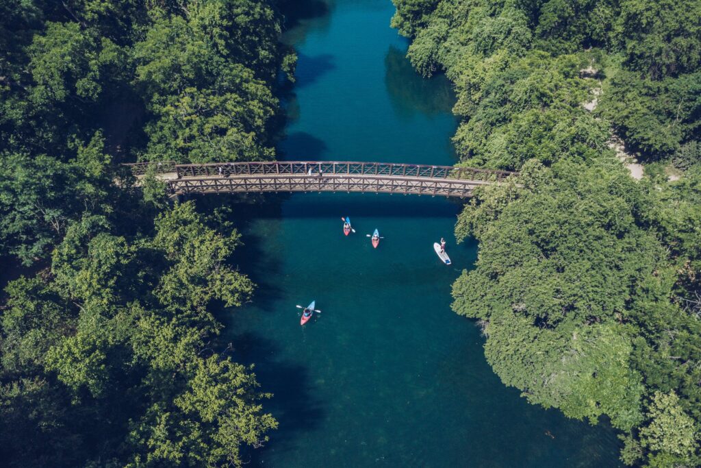Aerial view of kayakers on a river under a wooden bridge surrounded by lush greenery