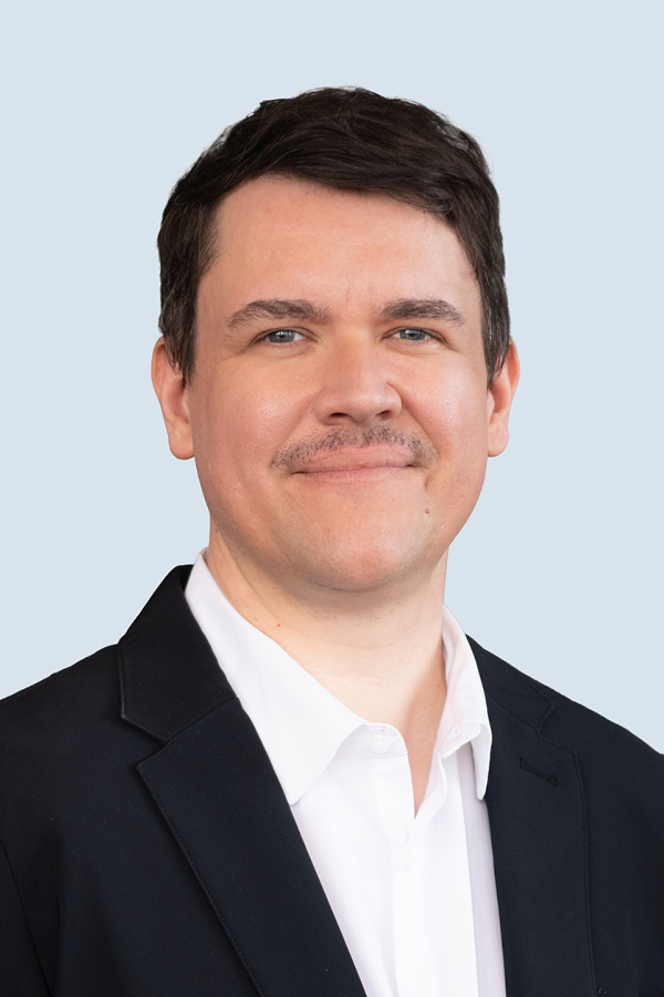 Smiling man in a black suit jacket and white pocket square against a plain background.