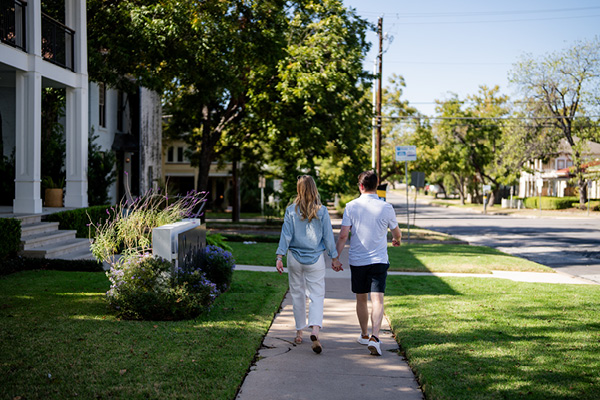 Woman and man walking hand in hand down a quiet residential street with green trees and lawns.